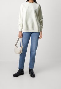 Donna che indossa un felpa bianca, jeans blu, stivali neri alla caviglia, mentre tiene in mano una piccola borsa beige su uno sfondo grigio uniforme.