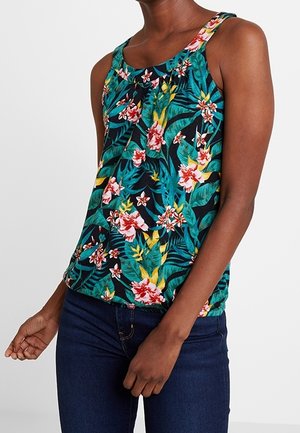 Ærmeløs tropisk blomstret bluse i grøn, gul og rød båret af en person med mørkeblå jeans mod en ensfarvet hvid baggrund.