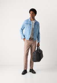 Veste en denim bleu clair, t-shirt gris, pantalon en lin beige et chaussures en cuir noires. Portant un sac de sport noir avec des accents argentés.