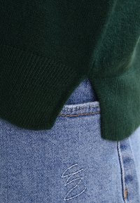 Ciemnozielony dzianinowy sweter o prążkowanej fakturze, z bocznym rozcięciem na dole, zestawiony z jasnoniebieskimi dżinsami z postrzępionymi detalami.