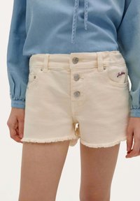 Shorts en denim beige clair avec un ourlet effiloché, fermés par trois boutons en métal argenté, dotés de poches avant et d'une broderie de marque discrète.