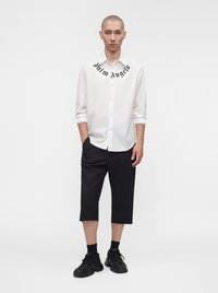 Mann trägt weißes Langarmhemd mit schwarzem Text am Kragen, schwarze knielange Shorts, schwarze Socken und schwarze Sneaker, steht vor weißem Hintergrund.