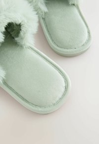 Lichtgroene pantoffels met zachte nepbont afwerking, zachte stoffen bovenzijde, platte zool en een ronde neus. Gestikte randen zorgen voor een gestructureerd detail.