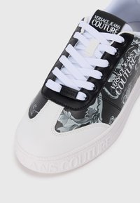 Bela teniska z črno-sivim cvetličnim vzorcem na boku, z belimi vezalkami, teksturiranim podplatom in poudarki logotipa znamke.