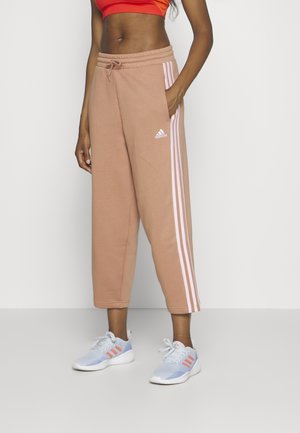 Vrouw draagt beige Adidas joggingbroek met witte strepen en lichtblauwe Adidas sneakers, staand met één hand in de zak.