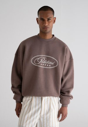 Sudadera marrón con cuello redondo y texto blanco "Prohibido", con una tela suave y texturizada y hombros caídos, combinada con pantalones rayados.