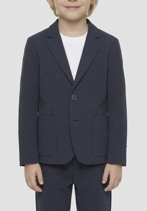 Enfant portant une veste de costume texturée bleu marine et un pantalon assorti sur une chemise blanche, debout devant un fond uni.