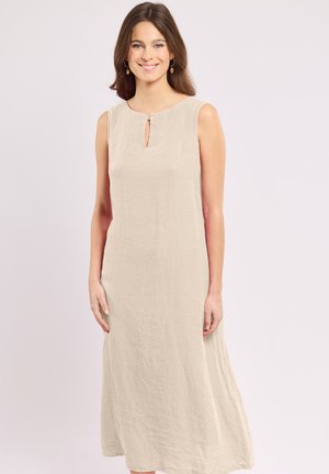 Femme souriante, debout, portant une robe en lin beige sans manches avec un petit col en forme de goutte et des boucles d'oreilles dorées pendantes sur un fond clair uni.
