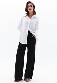 Camisa blanca de manga larga con cuello, combinada con pantalones holgados negros y zapatos marrones texturizados. Diseño simple, tejido suave, ajuste relajado.