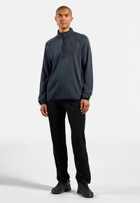 Pulloverjack met ritskraag in donkergrijs en zwarte borstzak; gecombineerd met zwarte broek en zwart schoeisel. Gladde textuur.