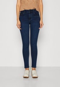 Hubené modré džíny s vysokým pasem, které mají dvě přední kapsy a hladkou texturu. Kombinované se světlými dřeváky.