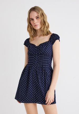 Blondynka w krótkiej granatowej sukience w białe groszki z bufkowatymi rękawami i guzikami z przodu, stojąca na jednolitym tle.