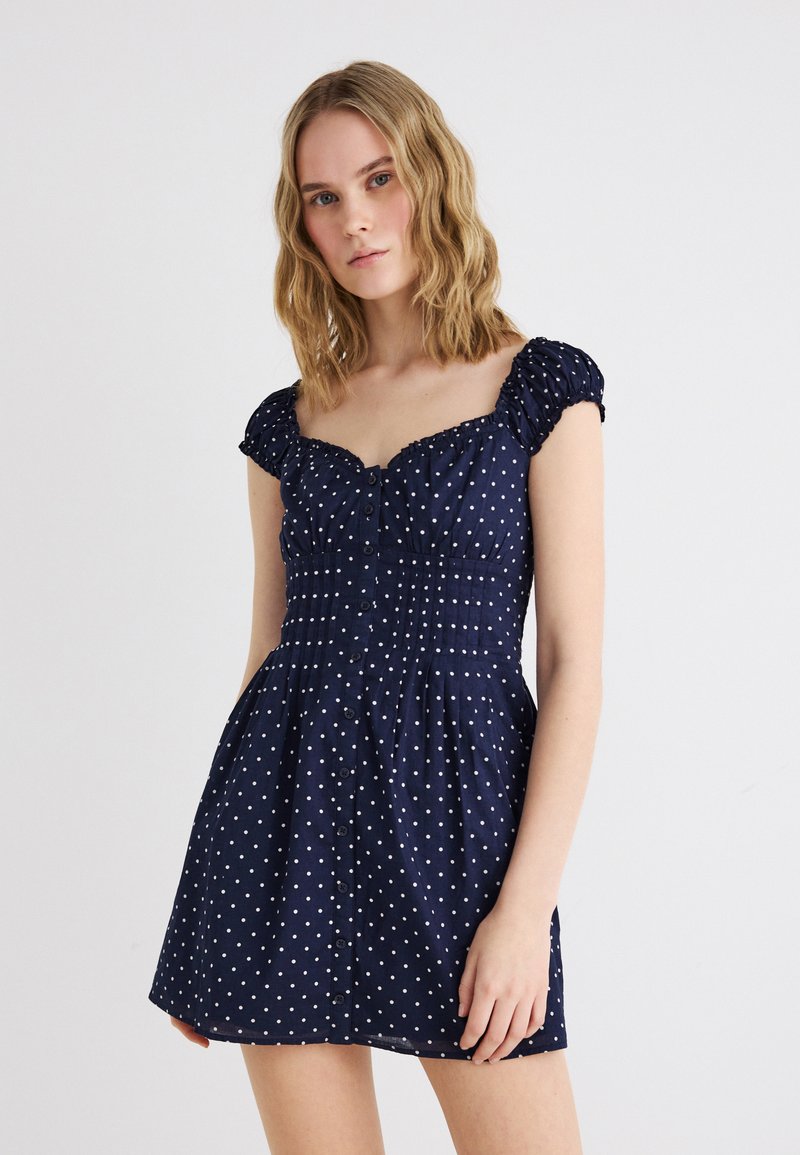 Ξανθιά γυναίκα που φοράει ένα κοντό φόρεμα σε σκούρο μπλε χρώμα με λευκές πουά, φουσκωτά μανίκια και κουμπιά στο μπροστινό μέρος, στέκεται σε απλό φόντο.