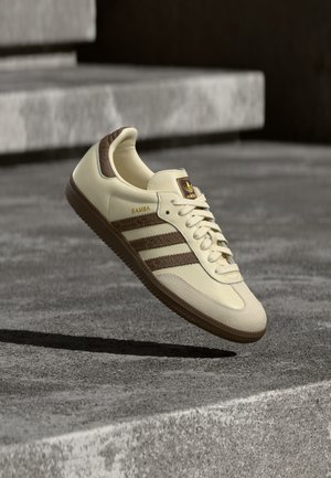 Crème en bruine Adidas Samba sneaker hangend boven betonnen trappen met branding zichtbaar aan de zijkant en op de tong.