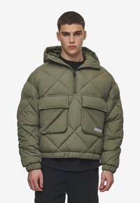 Giacca puffer trapuntata verde oliva con cappuccio, dotata di due tasche frontali, chiusura a mezza zip e polsini elastici sulle maniche.