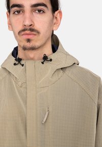 Chaqueta ligera beige con un patrón de cuadros, que cuenta con capucha, cordones ajustables y un bolsillo con cremallera en la parte frontal.