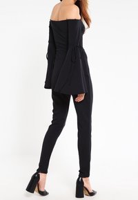 Svart avtagbar axeltop med långa klockärmar, knutna vid ärmsluten. Matchad med slim-fit svarta byxor och glänsande klackskor.