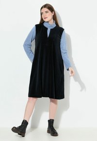 Schwarzes Cord-Kleid ohne Ärmel mit V-Ausschnitt und gefaltetem Rock, darüber getragen ein blau-weiß gestreifter Rollkragenpullover. Schwarze Schnürstiefel.