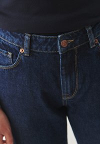 Jeans de mezclilla azul oscuro con un corte clásico. Las características incluyen un botón de cobre, costuras sutiles y un diseño estándar de cinco bolsillos.
