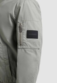 Manche de veste gris clair avec une poche zippée verticale et un patch rectangulaire noir affichant le logo "KHUJO&CO".