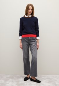 Maglione blu navy con orlo rosso sopra una camicia bianca con colletto, abbinato a jeans grigi a gamba larga e scarpe nere senza lacci. Texture liscia.