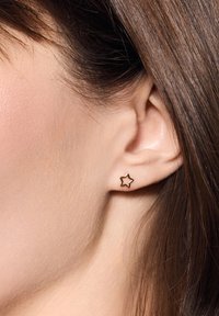 Złota gwiazdka w kształcie kolczyka, minimalistyczny design z otwartym konturem. Powierzchnia gładka i odbijająca światło, na tle jasnej karnacji.