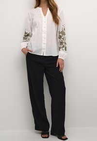 Blusa bianca di lino con bottoni davanti, pieghe sottili e ricami floreali sulle maniche; abbinata a pantaloni neri larghi e sandali.