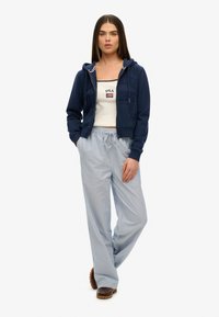 Hoodie navy avec zip, poches avant et lettrage ; débardeur blanc avec logo ; pantalons légers à rayures, coupe ample ; chaussures brunes avec semelles texturées.