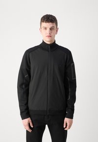 Black jacket with a high collar, featuring a zip front, textured fabric upper, and sleek lower section. Includes a side pocket with hardware.