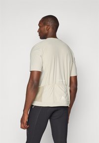 Ανοιχτό μπεζ τεχνικό t-shirt με πίσω τσέπη, κοντά μανίκια και ελεύθερη εφαρμογή. Διαθέτει μαλακή υφή και διακριτική σήμανση στον ποδόγυρο.