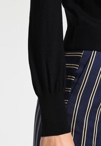 Svart stickad tröja med långa, puffiga ärmar. Närbild på ribbade muddar och ränder från marinblå och krämfärgade byxor. Mjuk textur.