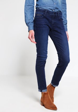 Osoba oblečená v tmavě modrých úzkých džínách s ohrnutými kotníky, v kombinaci s modrou denimovou košilí a hnědými semišovými kotníkovými botami.