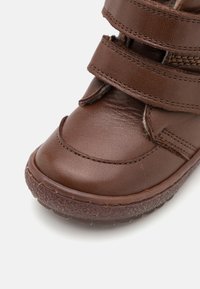 Botte en cuir marron avec trois sangles Velcro réglables, surface texturée, bout arrondi et semelle en caoutchouc robuste pour une bonne adhérence.