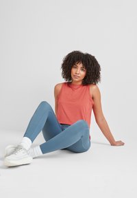 Woman with curly hair sitting on floor in blue leggings, coral sleeveless top, white sneakers, and socks against a plain light background.