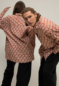 Camisas marrones con estampados y diseños geométricos blancos, de corte holgado, de manga larga, por fuera del pantalón. Llevadas con pantalones negros.