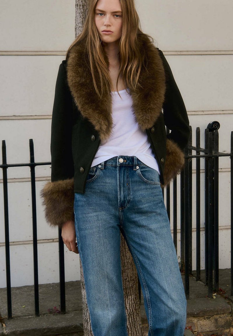 Jeune femme aux longs cheveux portant un manteau noir avec un grand col et des poignets en fourrure marron, une chemise blanche et un jean bleu, debout à l'extérieur.