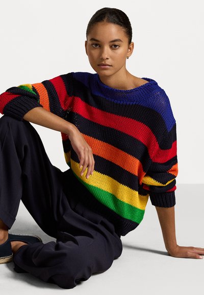 Pulover tricotat cu dungi multicolore (roșu, portocaliu, galben, verde, albastru, negru). Croială largă, decolteu larg, manșete din tricot. Pantaloni negri.