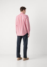 Camisa de manga larga en rosa con rayas verticales azules, de material de algodón, con cuello clásico, detalle de pliegue en la parte posterior, combinada con jeans oscuros y zapatos beige.