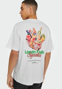 Witte katoenen t-shirt met een kleurrijke afbeelding van boodschappen in een draadmand. Inclusief de tekst "London Fields Superstore" in het groen.