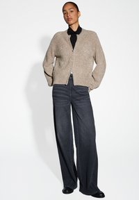 Vrouw in een beige sprankelende ritsvest over een zwarte coltrui, met een wijdvallende donkere jeans en zwarte schoenen, staande met haar handen in de zakken.