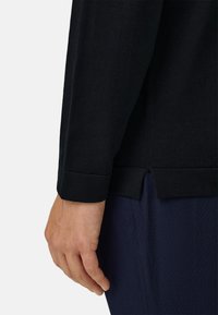 Main détendue sur le côté, portant une chemise à manches longues noire avec une fente latérale et un pantalon bleu foncé texturé sur un fond uni.