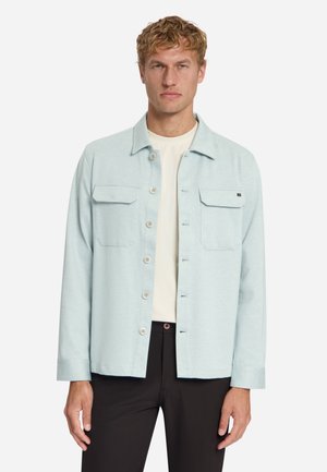 Mann trägt eine hellblaue Jacke mit Knöpfen über einem weißen Hemd und schwarzen Hosen und steht mit neutralem Gesichtsausdruck vor weißem Hintergrund.