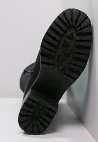 Suela de bota negra con ranuras profundas, material de goma, patrones angulares y una ligera curva en el talón; cuenta con una cremallera lateral para el cierre.