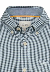 Koszula zapinana na guziki w niebieskie i białe kratkę. Posiada klasyczny kołnierzyk, beżowe guziki oraz małe logo na kieszeni na piersi.