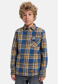 Chlapec s kučeravými vlasmi, oblečený v modro-horčicovožltom kockovanom košeli s gombíkmi a tmavosivých džínoch, stojaci proti jednému pozadiu.