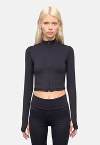 Schwarzes, kurzes langärmliges Sportoberteil mit hohem Kragen und Reißverschluss vorne, aus glattem, dehnbarem Stoff; kombiniert mit hochgeschnittenen Leggings.
