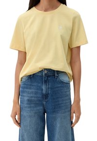Camiseta de algodón amarilla con mangas cortas, que presenta un pequeño logo de corazón blanco en el pecho, combinada con unos pantalones vaqueros de mezclilla azul de cintura alta.