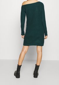 Abito lavorato a maglia off-shoulder di un verde tief. Maniche lunghe con texture a coste, lunghezza alla coscia e abbinato a stivali neri alla caviglia.