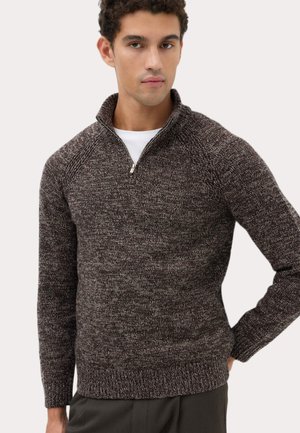Mand iført en brun og grå meleret halv-zip sweater over en hvid skjorte, stående med den ene hånd på hoften mod en ensfarvet baggrund.