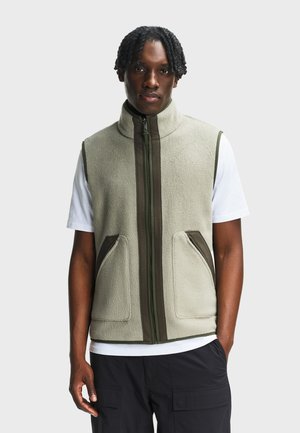 Mężczyzna z warkoczami, ubrany w jasnoszary polarowy bezrękawnik z ciemnozielonym obszyciem, na białej koszulce i czarne spodnie typu cargo, stoi z ręką w kieszeni.